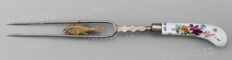 Two-pronged fork with a gilded plastic fish between the tines and a plastic palmette at the base; white metal fork, slightly curved white porcelain handle painted with colorful flower bouquets and insects on both sides. Ca. 1830, Korzec Korzec Porcelain and Faience Manufactory; 1790–1832. Two-pronged cutlery, height 30 cm., A metal two-pronged fork-like utensil with a curved narrow frame and a textured central fish-shaped decoration in gold tone, attached to a metal handle section that transitions into a white curved ceramic grip decorated with painted floral motifs in purple, red, pink, blue, green, and orange, set against a light gray background.