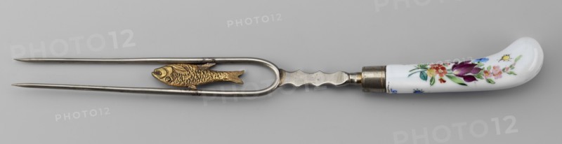 Two-pronged fork made of white metal with a gilded porcelain fish along the length between the tines and a porcelain palmette at the base. Slightly curved porcelain handle, white with colorful painted flower bouquets and insects on both sides. Ca. 1830. Korzec Korzec Porcelain and Faience Manufactory; 1790–1832. Cutlery; height 30 cm. Possible forgery., A metal two-pronged fork with a rounded U-shaped section near the handle holds a small decorative fish figure between the prongs; the metal is silver-colored and the fish is gold-colored with textured scales. The handle is curved and made of white material with painted floral decoration in colors including purple, red, pink, yellow, green, and blue, and there is a metal ferrule connecting the handle to the fork. The background is a neutral light gray.