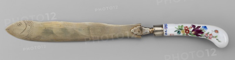 Knife, Korzec Porcelain and Faience Manufactory; 1790–1832. Possibly a Korzec forgery. Cutlery with a white-metal blade featuring a palmette at the base of the handle and an engraved fish at the end of the blade. Slightly curved porcelain handle with colorful painted flower bouquets and insects on both sides. Circa 1830. Poland Korzec?. Height 30 cm., A long flat metal blade with a rounded end engraved with a fish head and scale-like markings lies horizontally above a pale gray background, its metal showing a muted golden-silver patina with subtle uneven shading and a few small marks; the blade connects to a metal ferrule with a decorative leaf-shaped mounting and a short collar, which joins a curved glossy white handle painted with colorful floral motifs in shades of purple, red, pink, blue, green, and a small orange detail.
