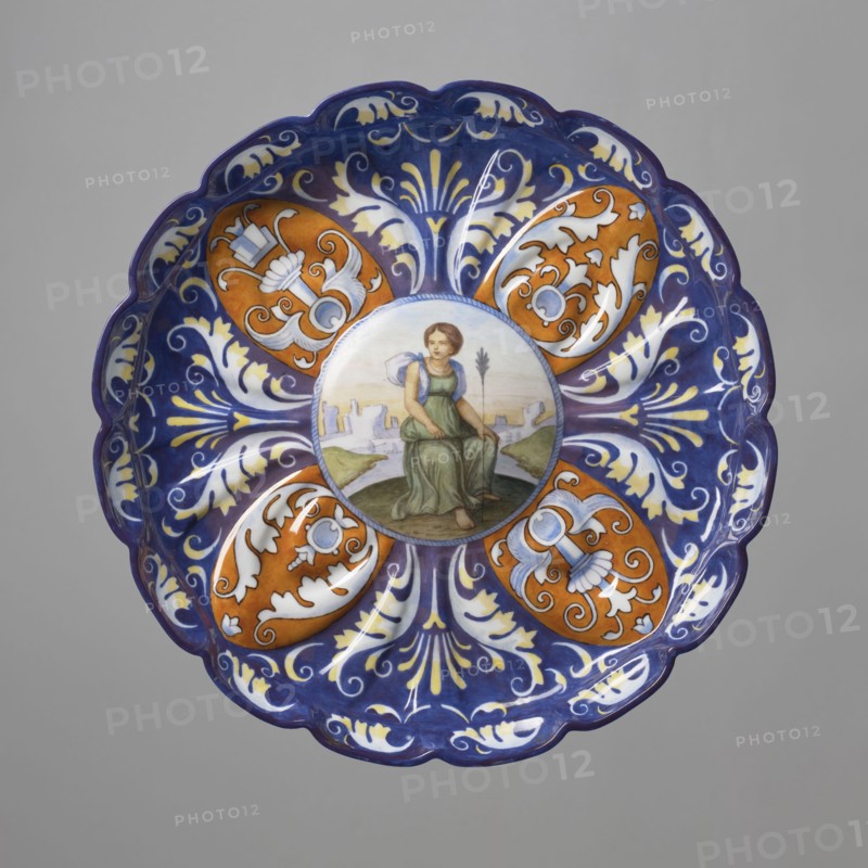 Patera-crespina, Koenigliche Porzellan Manufaktur KPM; Berlin; 1763–1918, imitation Italian majolica, crespina ceramics, Patera - crespisa. On a low hooped foot, modelled-fluted with many leaves, with a slightly raised multi-petaled edge and a raised-convex mirror. The mirror, bordered with a blue string motif, depicts a woman in a green tunic and blue shawl, sitting against a landscape with architecture, with a long arrow stuck in the ground in front of her. Around the collar, on a cobalt background, a motif of four ochre oval palmettes with inscribed decoration with blue plant-ornamental grotesque motifs, alternating with bipartite white-yellowish leafy grotesque motifs on a cobalt background; around the inner edge, a light leafy creeping ornament. Imitation of the form and decoration of 16th-century Italian maiolica a'quartieri from Faenza, 1849–1870, Germany, Berlin. Plateau; height 5.5 cm, width 30 cm., A decorative round plate with a scalloped edge, primarily blue and white with ...