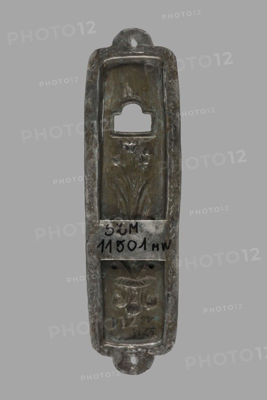 Mezuzah, Judaica; rectangular profiled sheet of low-grade silver edged with a flat ribbon decorated with a zigzag pattern and holes on the longer axis for nailing to a door frame; decorated with a bouquet of tulips in a vase, above it a rectangular opening with a semicircular finial; 1st half of the 19th century, Poland, height 10.1 cm, width 2.7 cm., A narrow vertical metal object with a rounded top and bottom and a central recessed panel, showing a worn metallic surface with areas of dark gray, brownish-green, and lighter silvery gray; the top recess has a rectangular opening with a rounded top edge, the middle recessed area features a floral relief with stems and petals, and the lower recessed area contains a decorative motif resembling a stylized vase and scrolls, all set against a plain gray background.