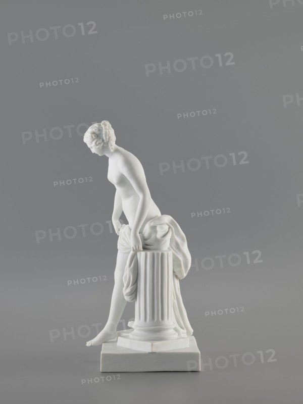 Venus Bathing, after Falconet; sculptor: Etienne Maurice Falconet 1716–1791 after Falconet's sculpture; manufacture: Churfuerstliche Porcellain Fabrique, Meissen 1763/74–1814. Figurine of Venus mythology, Marcolini's period 1774–1814. Porcelain sculpture/figurine, 18th/19th century, Meissen, Saxony. Depicts a young naked woman with long hair parted and arranged in a kind of crown; she stands in contrapposto by a fluted column cut at thigh height on a square plinth, slightly leaning forward, supporting with her hands the fabric falling from her left thigh, heavily draped and falling down the back along the column; she takes a step as if descending from a plinth onto a flat four-sided base. Height 25.4 cm; base width 10 cm; base length 12 cm., A white statue of a nude female figure leaning forward with her hands resting on a fluted cylindrical pedestal draped with cloth, the figure standing on a square base with one foot slightly extended, against a smooth gray background; visible colors...