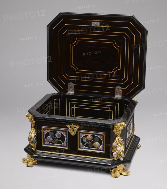 Jewel box, pietra dura technique; made of blackened wood, partly ebony; cuboid with chamfered corners supported on four gilded bronze volute legs decorated with a mascaron at the base. Edges of base and lid with profiled ebony strips. Decorated on side walls with six mosaic reserves and one plaque on the lid: front and back panels show two fruits on a branch; side panels show three fruits on a branch and an insect at the bottom. Corners feature cast gilded bronze garlands topped with medusa masks and plastic fruits made of semi-precious stones 7 pieces each. Hinged, profiled lid with a large plate showing three fruits on branches tied with a sash and an insect above; side walls decorated with gilded bronze garlands with plastic fruits; corners with gilded bronze shells; front with preserved keyhole. Dated to the early 18th century; attributed to Florence, Italy ; chest; height 34.5 cm, width 41.5 cm, depth 50.5 cm., A dark wooden octagonal chest with its lid open, showing concentric re...