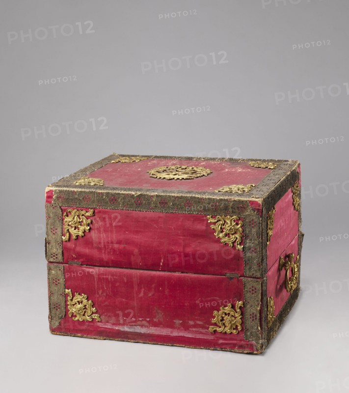 Casket case, gift, Maria Kazimiera Queen of Poland; 1641–1716, Strzalecki, Antoni Jan 1844–1934. The case is a rectangular wooden box covered with red velvet, with gold thread trimmings on the edges. The crosspieces have bronze openwork overlays. The side walls have gilded bronze handles; the top has a decorative central overlay with a rank crown. The front has a decorative overlay serving as a lock plate; the keyhole is preserved. The interior is lined with light green silk. Early 19th century; 18th century, Italy, Florence. Chest; height 39.5 cm, width 56.7 cm, depth 47.5 cm., A rectangular wooden box covered in red fabric with a worn surface and trimmed with dark textured edging; a group of ornate gold decorative metal appliques adorn the corners and center of the top and sides, and there are a few gold ring handles on the visible side; the box shows signs of wear with scuffs and faded areas on the red fabric, and the background is a smooth neutral gray.