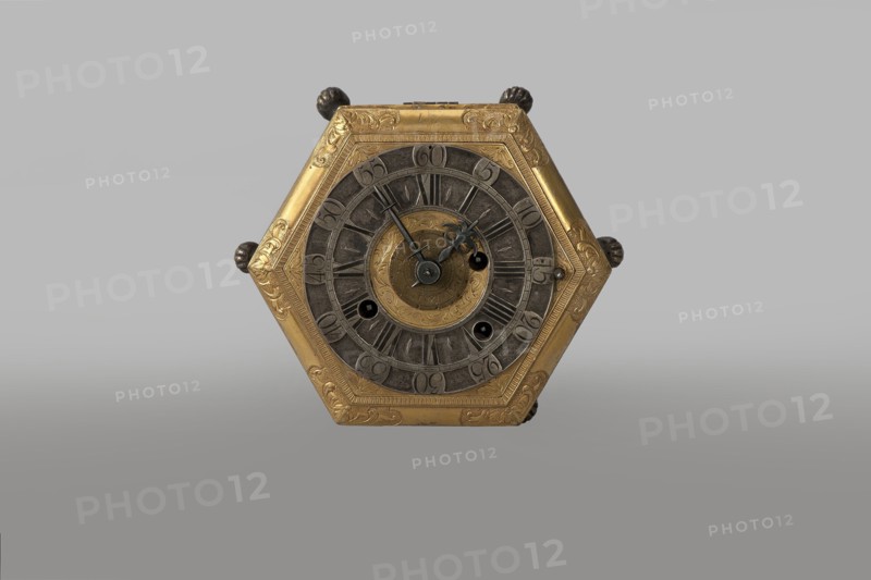 Tiled clock, Betzmeier, Erasmus fl. ca. 1750 clockmaker, acanthus ornament, lion's paws, mid-18th century, Warsaw, timepiece, height 8 cm, width 12.8 cm, A hexagonal metallic object with a central circular face is visible, showing a mix of gold and dark gray tones; the outer hexagon frame is gold with engraved decorative patterns, and the inner circular rings are dark gray with textured segments surrounding a small gold circular center that also has engraved designs; a group of dark bluish-gray hands and small circular openings appear on the central face, and small rounded dark knobs protrude from the hexagon edges; the background is a smooth gradient of light gray to medium gray.