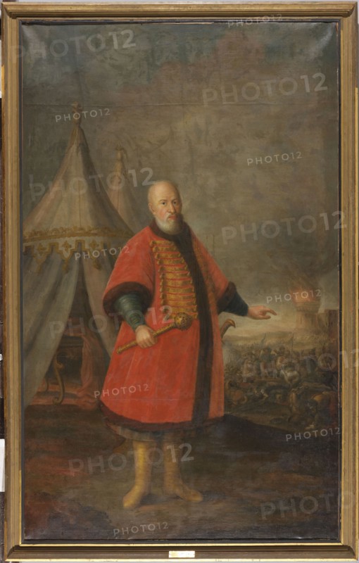 Portrait of Stefan Czarniecki 1599–1665, Crown Field Hetman, against the background of tents and a battle, unknown painter. Against the background of two round white-gray tents the first with a decorative lambrequin and a tuft on top, its flaps parted, in the dark interior a high table with S-shaped legs draped with red fabric. In the left foreground an old bald man with a short gray beard and mustache stands three-quarters to the right. He holds a baton in his right hand and with his left points to the battle raging in the background on the right. His face is oval with a high forehead; his blue eyes look directly at the viewer, his nose is prominent and slightly convex, his beard and mustache are closely cropped. He is dressed in a dark blue zupan with a red knee-length delia trimmed with brown fur at the neck, lapels, wide sleeves and hem, and wears high yellow leather boots. A saber hilt is visible at his left side. The battle scene shows a tangled crowd of horsemen before a bright ...