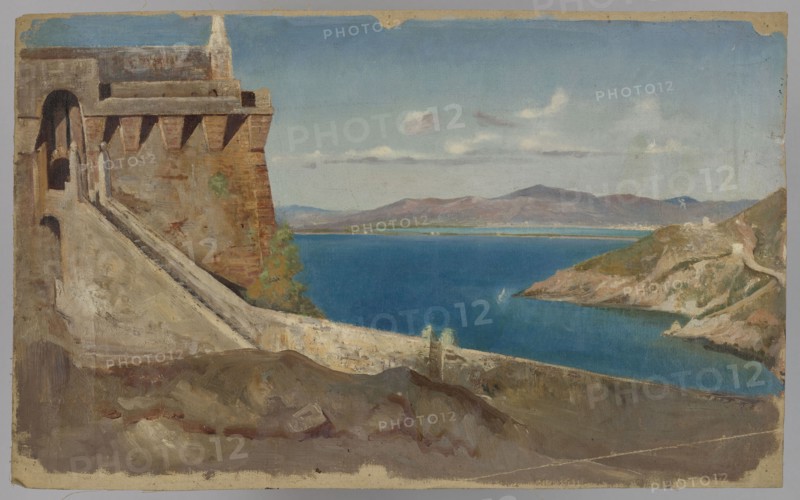Building by the Sea, Pius Welonski 1849–1931, painter; deposits, 1st quarter of the 20th century; painting; height 56 cm, width 34 cm, A coastal scene showing a large stone fortress or wall on the left with an archway and sloping ramp, rendered in brown and beige tones; a rocky foreground in muted browns and grays; a vivid blue body of water stretching across the middle; a rocky headland on the right with earthy browns, greens, and beige cliffs; distant low mountains in washed purples and browns beneath a pale blue sky with a few soft white clouds; small areas of green vegetation near the wall and along the shoreline.