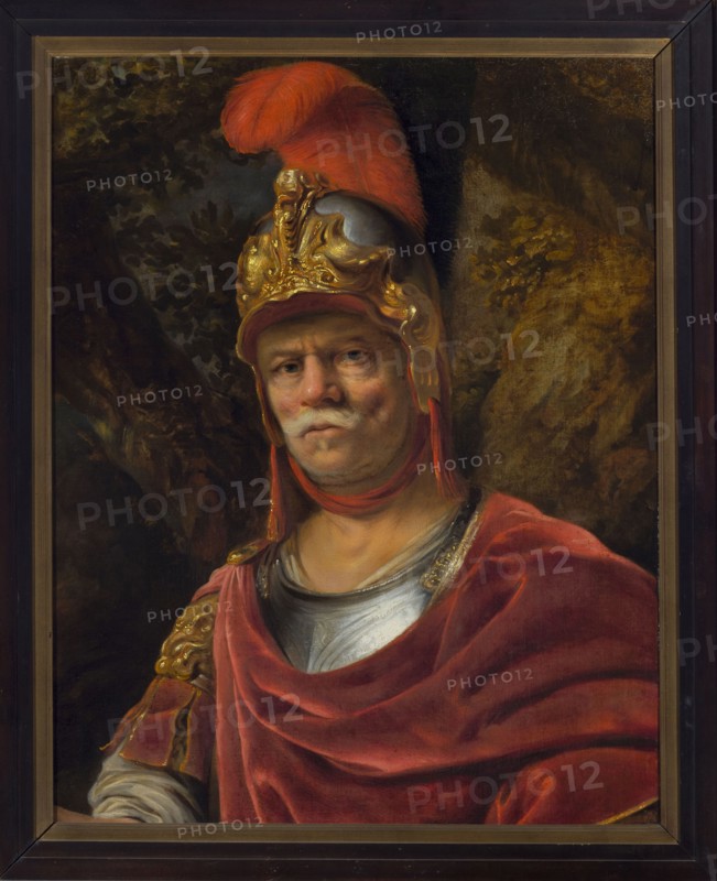 Man in a Golden Helmet Mars, Bol, Ferdinand 1616-1680 painter; armor; male portrait; helmet; Dutch painting; Stanislaw August Poniatowski King of Poland; 1732–1798; 1655; painting; height 72.5 cm; width 62.5 cm, A framed oil painting of an older man wearing a decorative metal helmet topped with a large red plume, with visible gold ornamentation on the helmet; he has a pale face with a white mustache and a neutral expression, and he wears a red cloak draped over armor with a silver breastplate and gold-accented shoulder piece; the background shows dark, leafy foliage and rocky textures in muted browns and greens, and the frame is dark brown with a lighter inner trim.