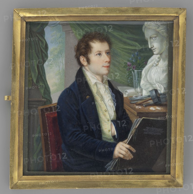 Portrait of a young sculptor with a sketchbook, pendant to Min.48. Painter: Schalck, Heinrich Franz 1791–1832. Interiors atelier; male portrait, half-length, sitting, 3/4 right. Young man with dark hair à la Tite, sideburns, wearing a white shirt and jabot, white vest with green stripes, navy blue frock coat with black collar and gold buttons. In his right hand a black sketchbook inscribed Skitzen. Background: left a column with a green curtain, right a table; above a plaster bust of a woman, beside it sculpting tools, a glass bottle with roses, forget-me-nots and bells. Medium: miniature with gilded bronze mounting. Date: 1820. Dimensions: height 9.0 cm, width 8.3 cm. Location: Wroclaw., A seated young man with fair skin and short brown hair wears a dark blue coat and a light striped waistcoat with a white ruffled shirt, holding a dark book and a rolled object in his hands; he faces slightly to the right with a neutral expression. Behind him is a draped green curtain and a pale column...