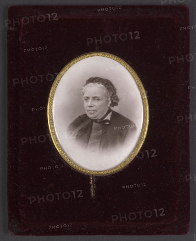 Portrait of Mlle Rousset at about age 70 died 1893, secretary and librarian of Izabella; photographer: Braun, Adolphe 1812–1877 from 1889 Braun, Clément et Cie, and 1910 – Braun e. Inscription notes: Josephine Rousset, niece of the Czartoryski doctor younger than Izabella, a student of the Hotel Lambert, a companion of the Czartoryskis' youth, over time gathered in her hands the administrative threads of Izabella's activities, and partly of Wladyslaw Czartoryski's. Date: 1870, place of creation: France. Medium: miniature. Dimensions: height 6.8 cm, width 5.4 cm. Subjects/attributes: en face portrait, woman, headgear., A small oval framed portrait of an older person with light-colored hair and a dark head covering, shown from the shoulders up, set against a white oval mat and surrounded by a thin decorative gold rim; the portrait rests on a dark burgundy velvet background with visible texture and a narrow dark gray border around the outer edge, and the portrait shows tones of black, whi...