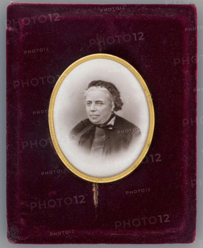 Portrait of Mlle Rousset at about age 70 died 1893, secretary and librarian of Izabella; photographer: Braun, Adolphe 1812–1877 author; from 1889 Braun, Clément et Cie, and 1910 – Braun e; inscription/note: Josephine Rousset, niece of the Czartoryski doctor younger than Izabella, a student of the Hotel Lambert, a companion of the Czartoryskis' youth, over time gathered in her hands the administrative threads of Izabella's activities, and partly of Wladyslaw Czartoryski's.; dated 1870; place of creation: France; miniature; height 6.8 cm, width 5.4 cm; en face portrait of a woman wearing headgear., An oval miniature portrait of an older person with light skin and short hair, wearing a dark garment and a dark head covering, set within a thin gold frame; the portrait sits against a textured deep burgundy velvet background with visible wear and small fiber details, and a narrow strip of gold beneath the frame.