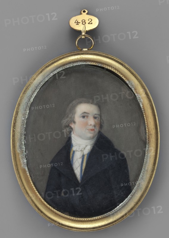 Portrait of a Man in a Black Frock Coat, Legrand, Franz 1807–1833 painter. Subject: men's portrait, 3/4 right, to the waist; young man with ash-grey hair and grey eyes. Costume: white cravat, vest with blue and white stripes and white checkered front, black frock coat. Medium/type: miniature. Binding: oval rim made of gilded brass, cylindrical, with a braided motif in the center. Date/place: 1850, Germany. Size: height 8.4 cm, width 6.7 cm., An oval portrait set in a gold-colored metal frame with a small loop and hanging tag at the top shows a man with light skin and shoulder-length brown hair wearing a dark coat and a white cravat; his face has pale skin with pinkish cheeks and he looks slightly to one side against a muted gray background, with visible wear and discoloration along the inner edge of the frame and a few blue and yellow accents on the man's clothing.