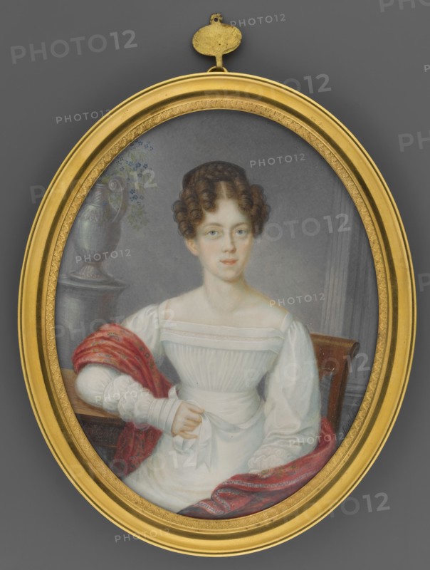 Portrait of a Woman in a White Dress Sitting in an Armchair, by Heinrich Franz Schalck 1791–1832, oil miniature, 1812. Subject: woman en face in an interior with flowers in a vase; costume and hairstyle of the Empire period Empire dress, low neckline. Miniature, height 13.6 cm, width 10.8 cm., An oval framed portrait shows a seated young woman with fair skin and brown curly hair styled close to her head, wearing a white dress with puffed sleeves and a fitted bodice; she has a calm facial expression and is posed with one hand holding a white sash at her waist and the other resting on a surface draped with a red patterned shawl. The background includes a muted gray wall and a dark wooden chair, and a decorative urn with a few small blue flowers sits on a pedestal beside her. The frame around the portrait is gold, and visible colors in the image include white, brown, red, gold, gray, blue, and muted green.