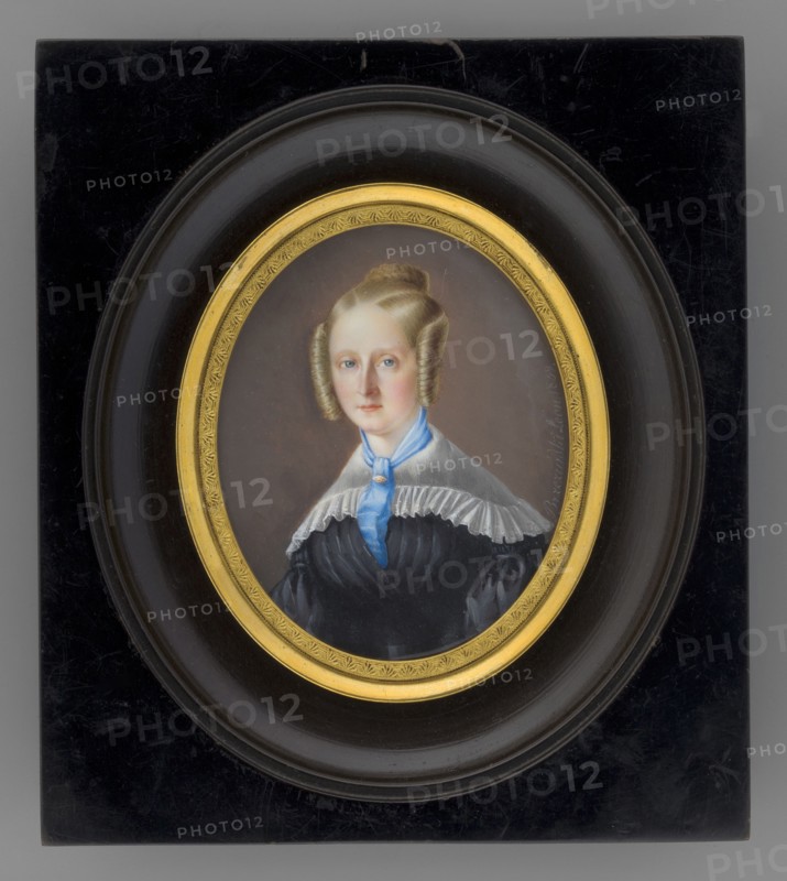 Portrait of Mrs. Ordezyna? Antonina née Kielczewski 1811–1893, wife of Józef Ordega, Lodzia coat of arms 1802–1879. Painted by Leon Brzezinski 1809–1865. Female portrait, three-quarters, young woman, half-length, almost straight posture, light blond hair in spiral curls along the face, braided crown at the top of the head. At the neck a blue cravat fastened with a ruby brooch; white gauze cape-like collar on the shoulders; black dress with a wide belt at the waist. Background in tones of broken brown. Oval miniature, signed “Brzezinski Leon, 1839.” Inscription on the reverse: “p. Ordega”, 1839, France. Miniature set in an oval gilded-bronze frame with palmette motif within a polished black rectangular wooden frame under convex glass. Dimensions: height 7.4 cm, width 6.0 cm., An oval portrait of a woman set in a rectangular dark frame with a rounded inner border and a gilded inner ring; the woman has light skin and light brown hair styled in symmetrical rolled curls at the sides and a b...