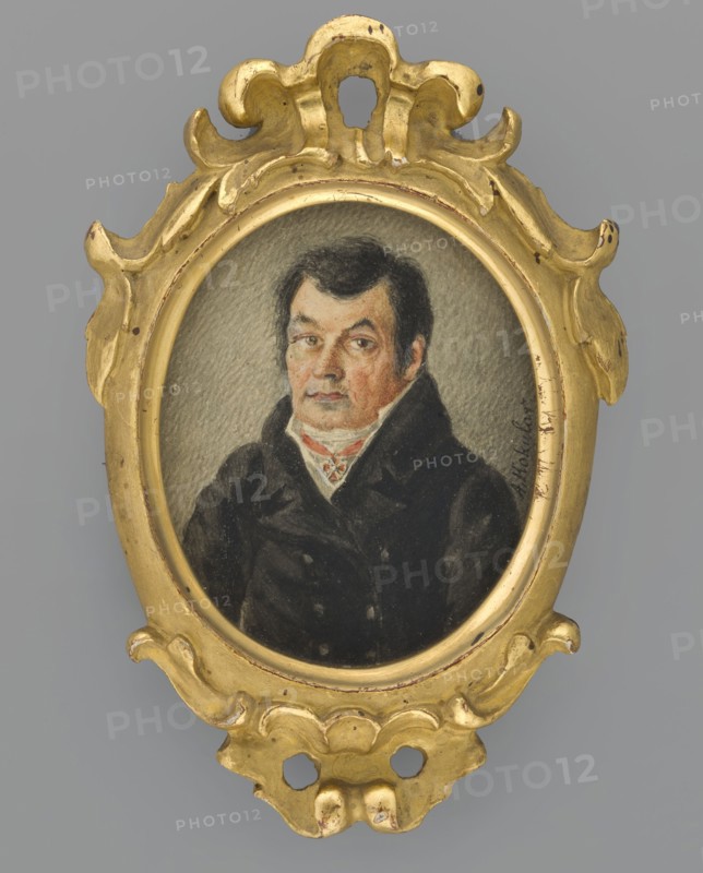 Portrait of a Man in a Black Frock Coat, Kokular, Aleksander 1793–1846, painter. Portrait of a man, half-length, three-quarters left. Short black hair; white cravat, white vest, black double-breasted frock coat; wearing the Order of St. Anne, 2nd Class. Background grey, lined. Oval wooden frame with Baroque ornament, plastered and gilded. Inscribed on front: A. Kokular. On back of frame: Pal. Kokular Alek. / Miniature: 'portrait of a man' / Polish painting / Catalog No. 20 / Miniature / Poland. Dated 1840. Miniature, height 6.3 cm, width 5.1 cm., An oval portrait of a man set in an ornate carved frame, the frame painted gold with scrolled and rounded decorative shapes and small areas of wear showing a darker base; the portrait shows a man with dark hair and sideburns wearing a dark coat and a light-colored high collar with a patterned cravat, his face rendered with flesh tones and subtle shading against a neutral gray background; visible colors include gold, black, dark brown, gray, be...