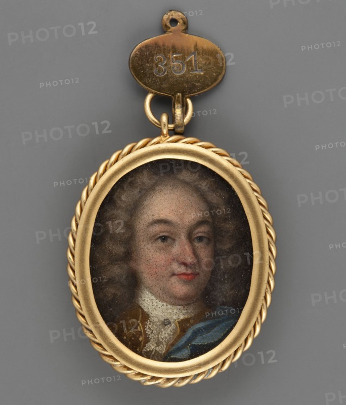 Portrait of a man in a lion-style wig, Georg von Hessen-Darmstadt 1669–1705 ?, unknown artist, men's portrait 3/4 right, aristocracy, costume tailcoat, jabot, hairstyle lion-style, jewelry brooch at the jabot, foreign miniature, 1750, miniature, height 3.4 cm, width 2.8 cm, An oval pendant with a twisted gold frame enclosing a painted portrait of a person with pale skin, flushed cheeks, light brown curly hair, and a neutral expression, wearing a white lace collar and garments with brown and blue tones; the pendant is attached by gold rings to a small oval gold tag, and the background behind the pendant is gray.