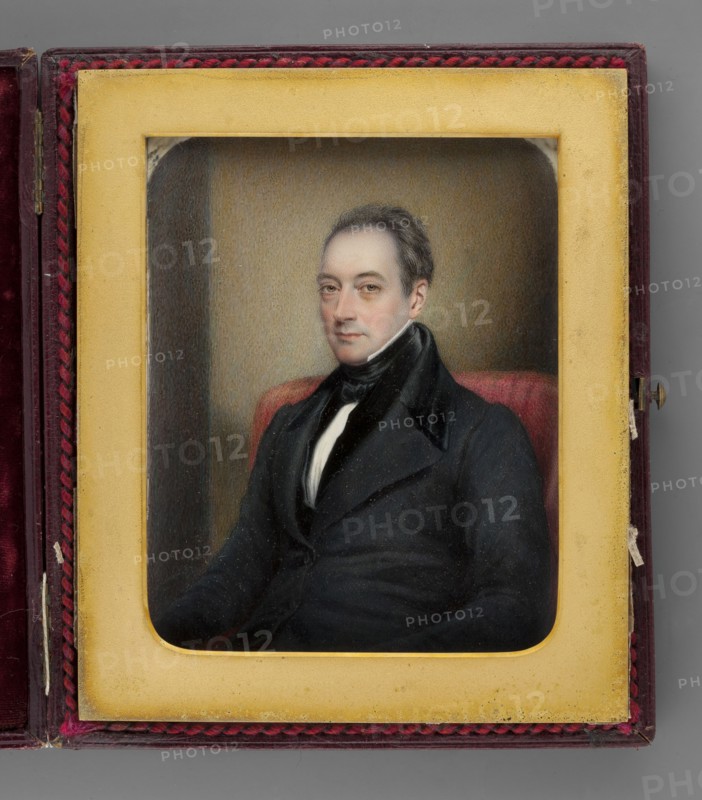 Portrait of a Man in a Frock Coat, painted by Henry Collen 1800–fl.1872, 1837. Seated waist-high, three-quarter left. Dark salt-and-pepper hair, hazel eyes. Wearing a white shirt, tie, vest, and black frock coat. Background shows the back of an armchair with red upholstery and a yellow-brown-gray wall. Miniature on cardboard, height 12.1 cm, width 9.7 cm. Binding: cherry leather case, metal frame with braided border, passe-partout. Inscribed bottom left: “H. Collen / 1837.” On the reverse in ink: “1837 / Painted by Henry Collen / miniature painter to / the Queen / and HRH The Duchess of Kent / 37 Somerset Street / Portman Square / London.” Pencil inscription on the cardboard edge: “Col Porter Cate / XII, 1837.”, A painted portrait of a seated man displayed inside a rectangular frame and a hinged case; the man wears a dark coat with a high collar and a white shirt visible at the neck, and he has short dark hair and a neutral expression. The frame around the portrait is a yellowish-cream...