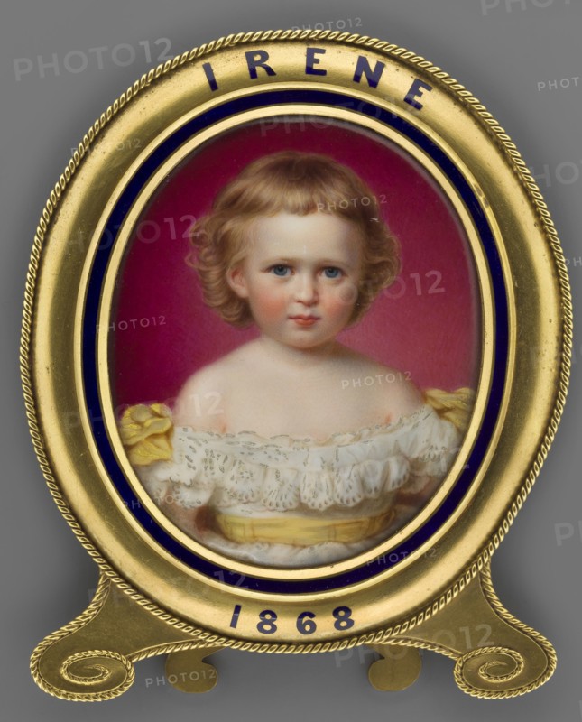 Irene von Hesse und bei Rhein 1866–1953, daughter of Louis IV of Hesse, portrayed as a girl; after a miniature by Reginald Easton 1807–1893. Portrait miniature, England, 1868. Subjects: children, girls, rulers. Medium: miniature; dimensions: height 8.1 cm, width 6.7 cm., An oval framed portrait of a young child with light brown curly hair and blue eyes, wearing a white lace dress with yellow ribbon details, set against a deep pink background; the portrait is enclosed in a gold frame with dark blue accents and a decorative rope-like edge, all shown on a gray surface.