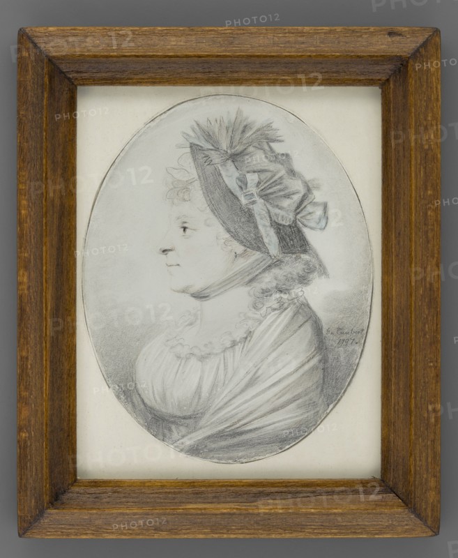 Portrait of an Elderly Woman Wearing a Hat in Profile, Taubert, Gustav Friedrich Amalius 1755-1839 painter. Women older, hats, headgear, profile left, female portrait, foreign miniature. Torso of an elderly woman in left profile. Hair in small curls revealing the forehead and falling to the shoulders. The hat is trimmed with a pale blue ribbon tied under the chin. The dress is high-waisted; the oval neckline is decorated with a ruffle, and a shawl with a faint bluish shadow is worn on the shoulders. Background: natural color of the paper. Binding: secondary, contemporary, wooden, 1797, Gdansk. Miniature, height 8.7 cm, width 6.9 cm., A framed oval portrait drawing of a woman shown in profile facing left, rendered in shades of gray and black on a light background; she wears a bonnet with ribbons and feather-like decoration, curly hair visible at the nape, a high neckline with ruffled trim, and a draped garment, all depicted with delicate pencil or charcoal shading; the frame is wooden w...