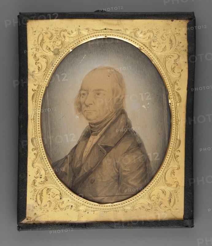 Cyprian Szukiewicz ca. 1775–1844, godfather of Cyprian Kamil Norwid; painter; portrait in three-quarters of a man; male portrait; Polish miniature; amateur artist; ; subject: man of letters; dated 1860; place of creation: Poland; miniature; 11.5 × 9 cm., An oval portrait of an older person with light skin tones and pale hair is set within an ornate rectangular frame; the person wears a dark coat and a high collar with layered garments visible, and their expression is neutral. The portrait background is a muted grayish-blue gradient. The frame surrounding the oval is gold with intricate floral and scroll embossing, and the outer border of the rectangular case is black. Overall visible colors include pale skin tones, light beige, grayish-blue, brown, gold, black, and muted dark brown.