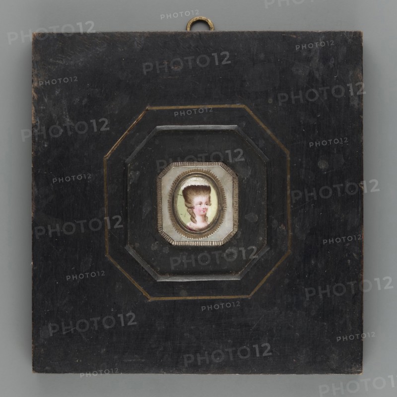 Woman's head in a high wig. Portrait en trois-quarts of a woman with a high blond wig trimmed with a white headdress at the top; white trim on a magenta dress at the bodice. Background: yellowish-green. Oval bronze frame set in an octagonal mother-of-pearl plate placed in an octagonal cut of a square black unpolished wooden frame, inlaid with bronze strips. Miniature, dated 1775, height 1.5 cm, width 1.2 cm., Polish miniature; artist possibly Leopold Méyet 1850–1912 noted in association; amateurs;, mentioned., A square dark wooden panel with worn surfaces and a small metal hanging loop at the top surrounds a recessed octagonal frame with layered stepped edges and subtle gold trim; set in the center is a tiny painted portrait of a person with pale skin, high powdered brown hair, pink cheeks and lips, and a visible white cap, all within a small decorative bezel, with colors including black, dark brown, gold, white, pink, brown, beige, and pale green.