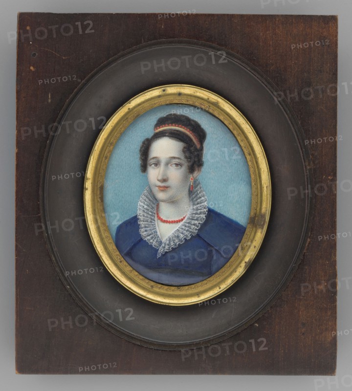 Portrait of a woman in a blue dress with a ruff, unknown artist, three-quarter portrait, costume, female portrait, Polish miniature, amateur, dated 1815–1830, miniature, height 5.6 cm, width 4.8 cm, A framed oval portrait of a woman set in a dark brown wooden plaque with a recessed black circular mat and a gold inner frame; the woman has dark brown hair styled up with a red band, pale skin, and neutral facial expression, wearing a blue dress with a high white lace ruffled collar and a red bead necklace, with a single red drop earring visible, and a light blue background behind her.