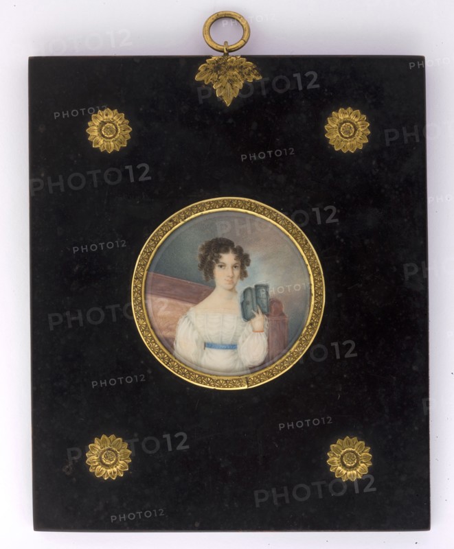 Portrait of a woman reading a book, unknown author amateur; books; woman; full-face portrait; interior; female portrait; hairstyle; Polish miniature; amateur; 1825; miniature, A rectangular black plaque with a glossy finish features a central round miniature portrait set in a decorative gold frame; the portrait shows a person with curly dark brown hair wearing a white dress with a blue sash and holding a small blue-gray object, seated against a muted background of soft gray and brown tones. The plaque is embellished with several ornate gold floral or sunburst motifs near the corners and a gold loop and leaf-shaped ornament at the top. The visible colors include black, gold, white, blue, brown, gray, and subtle flesh tones.
