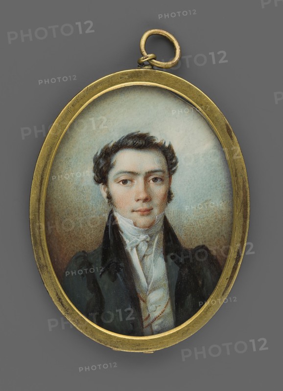 Portrait of a young man in a black tailcoat, en face portrait, male, nobility, Polish miniature, unknown artist, 1826, miniature; height 5.2 cm, width 4.0 cm., An oval pendant with a gold-colored metal frame and loop contains a painted miniature portrait of a young man shown from the chest up; he has dark brown wavy hair, fair skin with a slight rosy tint, and dark eyes with a neutral expression. He wears a high-collared white shirt and cravat beneath a dark coat with broad lapels, and the background inside the pendant is a soft, muted mix of pale blue and beige tones while the outer area around the pendant is a medium gray.