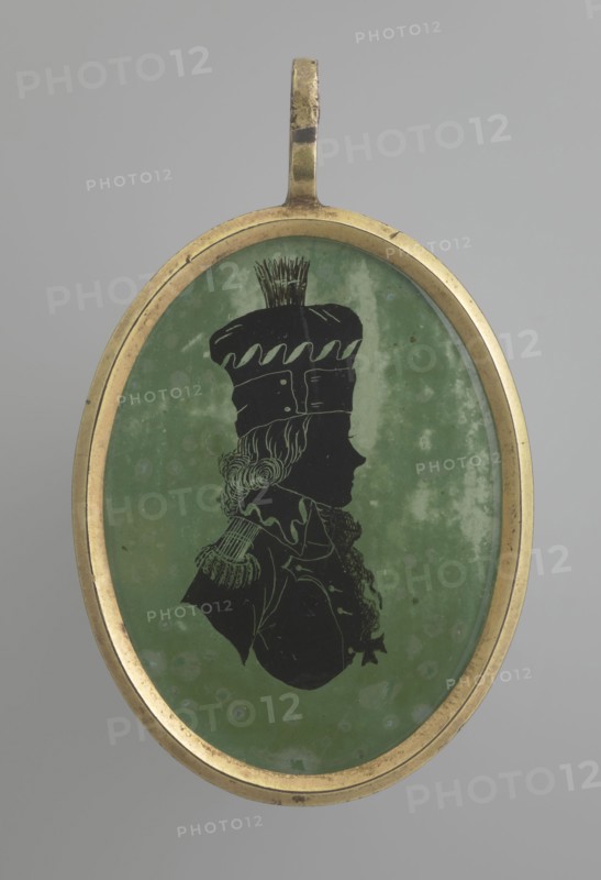 Tadeusz Kosciuszko in a general's uniform, profile silhouette. Black silhouette on a light green background showing Kosciuszko's right profile with mid-long hair emerging from under a high cap with a brim and a ponytail on the crown; half-bust in uniform with epaulettes and a high collar, a scarf or ribbon at the neck from which a cross-order hangs. Background with lighter spots and abrasions to the right and above the head. Oval brass frame. Miniature, circa 1800, height 4.4 cm, width 3.2 cm., An oval pendant with a gold-colored metal frame containing a silhouette portrait on a mottled green background; the silhouette is black and depicts a person in profile wearing a tall hat with decorative top details and a garment with a high collar and textured shoulder decoration; the frame shows slight wear and the overall colors visible are gold, green, black, and shades of gray in the background.