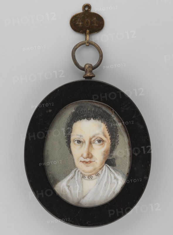 Portrait of an Elderly Woman in a Bonnet, Unknown artist. Subject: elderly woman, shoulder-length portrait, head slightly turned left. Wearing a black bonnet, neck band embroidered with black and white dots, white dress. Hazel eyes. Style: miniature painting, bust almost full-face. Date: 1880. Materials/format: flat oval wooden frame; miniature, height 4.8 cm, width 8.9 cm., An oval pendant with a black outer frame contains a small painted portrait of a woman shown from the shoulders up; she has pale skin, dark curly hair pulled back, light-colored eyes with visible lashes, and a neutral expression with slightly pursed lips. She wears a white garment and a delicate choker-style necklace, and the painted background behind her is a muted gray with a stippled texture. Attached to the top of the frame is a metal ring and a small metal tag; the ring and tag show a worn metallic patina in brass and bronze tones, and the overall color palette includes black, white, pale skin tones, dark brown...