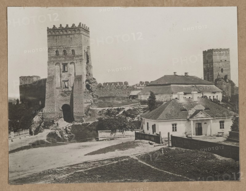 Lutsk. The Castle – view towards the Styrowa Swidrygiello Tower, /photographer, architecture, Polish architecture, Lutsk Ukraine, castles, ca. 1918 negative, ca. 1918–ca. 1922 print, Lutsk, photography, nature photography, photographic print: height 11.2 cm, width 14.6 cm; cardboard: height 35.1 cm, width 25.2 cm., The image shows an outdoor scene with a large stone tower ruin and sections of a fortified wall alongside a group of low buildings with pitched roofs; the tower and wall appear in shades of gray and brown, and the buildings are in lighter gray and off-white tones with darker gray roofing. In the foreground there is an unpaved area and a grassy patch in muted greenish-brown, bordered by a dark wooden fence and a few scattered shrubs and small trees in dark green. A path and a simple wooden gate lead toward the base of the tower, and there are several trees and low vegetation between the structures. The sky is pale and almost white, creating a high-contrast backdrop to the arc...