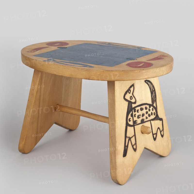 Table from a set of doll furniture. Wooden table with an oval top resting on stringer supports, connected with a lower link. Painted decoration: schematic silhouettes of animals on supports and tableware arrangement on the tabletop. Toy. Maker: Kenar, Antoni 1906–1959, sculptor; co-creators: State Secondary School of Fine Arts in Zakopane Zakopane; 1876–, Office of Production Aesthetics Supervision Warsaw; 1947–1949. Date: 1947–1948. Place of origin: Zakopane. Dimensions: height 12 cm, width 18 cm, depth 15 cm., A small wooden stool with a rounded top and two triangular legs joined by a dowel, the wood showing a light tan color with visible grain; the top surface has a central rectangular area painted blue and a few circular and elongated painted shapes in red and blue near the edges; one leg features a simple black line drawing of an animal with a spotted body and a raised tail; the background is a plain light gray.
