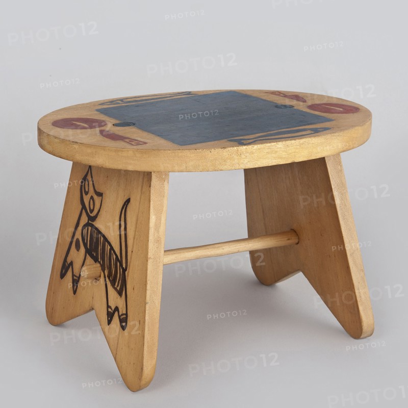 Table from a set of doll furniture, State Secondary School of Fine Arts in Zakopane Zakopane; 1876-, co-creator, Office of Production Aesthetics Supervision Warsaw; 1947-1949 co-creator, Kenar, Antoni 1906-1959 sculptor/author, Wooden table with an oval top resting on stringer supports, connected with a lower link. Painted decoration—schematic silhouettes of animals on supports and tableware arrangement on the tabletop. 1947–1948, Zakopane; toy. Dimensions: height 12 cm, width 18 cm, depth 15 cm., A small wooden stool with a round top and three angled legs joined by a horizontal dowel, showing light tan and beige wood tones with areas of darker brown grain; the top surface has a large dark blue painted rectangle and small circular and crescent-shaped motifs in red and blue, and one leg features a black hand-drawn animal-like figure with bold outlines and striped detail.
