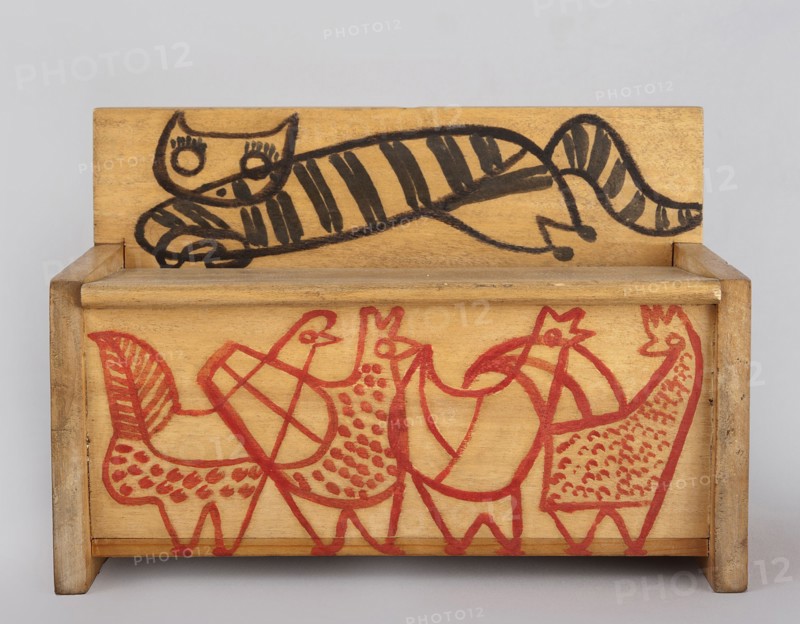 A wooden bench from a set of doll furniture. Wooden bench with a box closed by a flap forming a seat. Painted decoration with schematic animal silhouettes: a tabby cat on the backrest and chickens on the sides and front wall of the box. Toy, height 11.5 cm, width 17.0 cm, depth 8.0 cm. Maker: Kenar, Antoni 1906–1959, sculptor. Date: 1947–1948. Origin: Zakopane. Associated with State Secondary School of Fine Arts in Zakopane and Office of Production Aesthetics Supervision 1947–1949., A small wooden box with a hinged back shows a simple black line drawing of a reclining striped cat on the back panel and a group of stylized birds drawn in red across the front panel; the wood is a warm tan color with visible grain, the cat features bold black stripes and round eyes, and the birds are outlined and patterned with red marks.