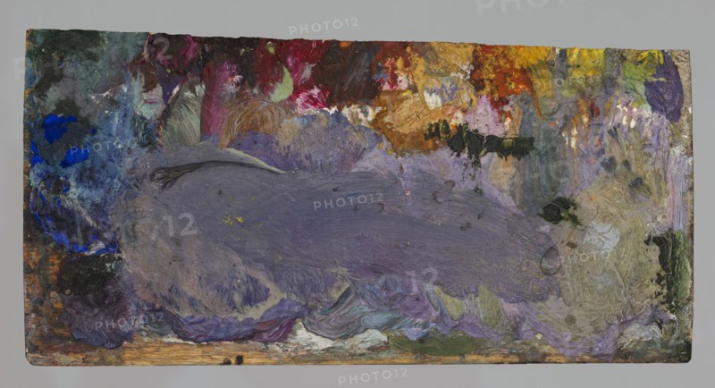 Jan Stanislawski's rectangular palette; Jan Stanislawski 1860–1907, painter; painting accessory — painter's palette; associated with plein-air painting and painter's workshop; late 19th century; varia; height 7.5 cm, width 14.4 cm, A rectangular surface covered in thick layered paint showing a large central area of muted purple and gray brushstrokes, surrounded by patches of vivid blue on the left, deep red and magenta near the top, warm yellow and orange toward the upper right, and areas of green and olive along the right edge and bottom; there are also creamy beige and off-white smudges, small dark black and charcoal spots, and a worn wooden strip visible along the bottom edge.