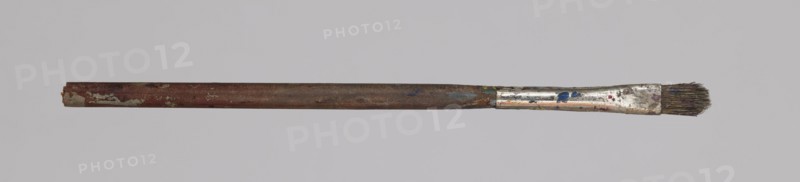 Jan Stanislawski's flat brush; Jan Stanislawski 1860–1907, painter. Painting accessory flat brush used in a painter's workshop and for plein-air painting, late 19th century. Dimensions: height 12.5 cm, width 0.5 cm., A single used paintbrush lies horizontally across a light gray background, with a long worn wooden handle showing reddish-brown and darker brown areas and spots of pale green paint, a metallic ferrule near one end that is scratched and mottled with silver and small splashes of blue and red, and a short cluster of dark bristles at the tip that appear stained and frayed.