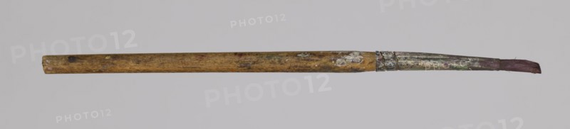 Jan Stanislawski's round brush; maker: Jan Stanislawski 1860–1907 painter; painting accessory: round brush for plein-air painting; late 19th century; dimensions: height 13.5 cm, width 0.7 cm; category: brushes, painting accessories, varia., A single used paintbrush lies horizontally against a pale gray background; the wooden handle is worn and mottled with stains in brown and yellowish tones, showing a few darker spots and areas of lighter discoloration, the metal ferrule is tarnished and splattered with remnants of dried paint in muted green, white, and silvery gray, and the bristles at the tip appear frayed and stained a dull reddish-brown.