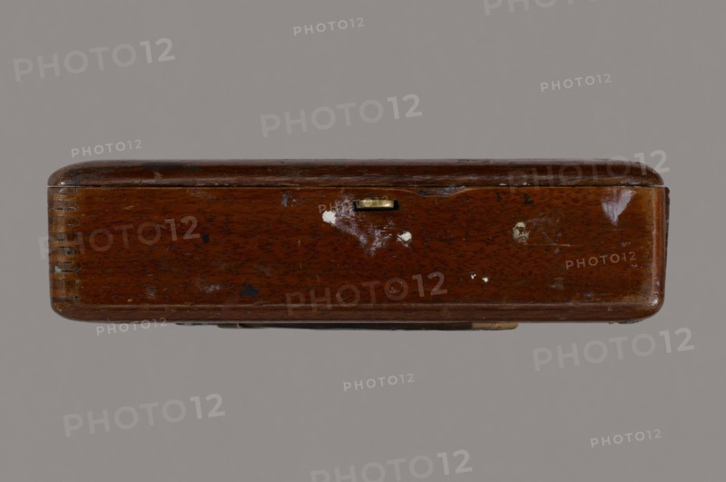 Jan Stanislawski's painting box; painter Jan Stanislawski 1860–1907; painting implements; artists in the open air; painting accessories; plein air studies; late 19th century; varia; height 10 cm, width 16 cm, depth 3.5 cm, A rectangular wooden box viewed from its long side with rounded corners and a glossy finish showing scratches and worn areas, a small metallic rectangular latch inset near the top center, a few small spots of chipped paint and scuffs across the surface, and a plain neutral background. The visible colors include brown on the wood, gold on the latch, white from the chipped spots, and a gray background.