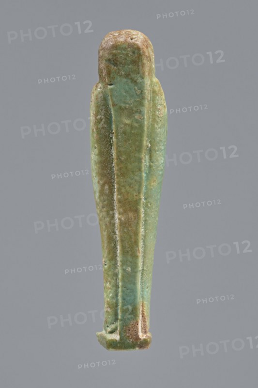 Ankhren's shabti, Ankh-ren name. Mummy-shaped figurine, funerary shabti, standing on a base and supported at the back by a pilaster. Arms crossed on the chest, holding hoes in both hands; a string in the right hand suspends a basket over the left shoulder. Convex tools. Smooth, three-part wig with ears uncovered. Facial details slightly blurred, large eyes, proportionate nose, straight long beard. Inscription engraved in a column on the front of the legs: The word spoken by Osiris Ankh-ren .... Date: 664–30 BC Late Period–Ptolemaic Period. Provenance: Tuna el-Gebel. Height: 10.8 cm., A vertical elongated figurine-like object with a rounded top and tapered bottom, shown against a plain gray background; the surface of the object displays a mix of green and turquoise hues with areas of brownish tan and pale beige patina, a textured and slightly worn finish with visible small pits and rough spots, and subtle linear ridges running vertically along its length creating narrow recessed panels ...
