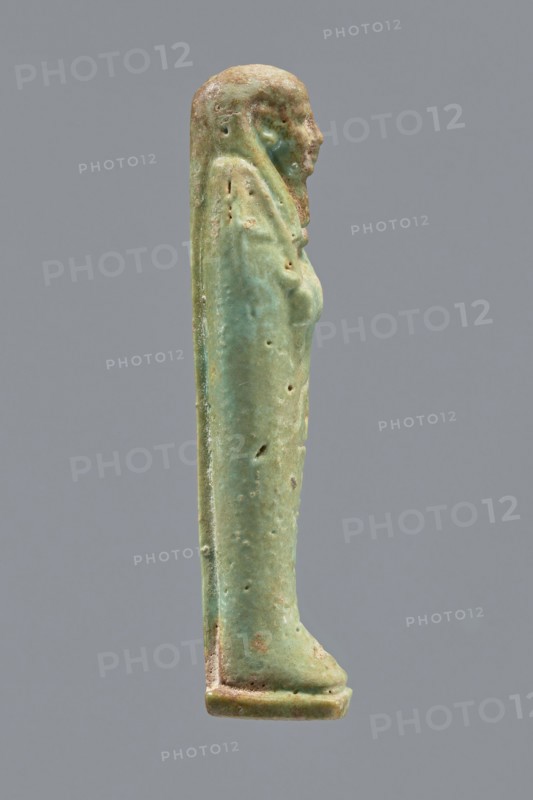 Ankhren's shabti, Ankh-ren name. Mummy-shaped figurine, funerary shabti, standing on a base and supported at the back by a pilaster. Arms crossed on the chest, holding hoes in both hands; in the right hand also a string suspending a basket over the left shoulder. Convex tools. Smooth three-part wig, ears uncovered, facial details slightly blurred with large eyes, proportionate nose, straight long beard. Inscription engraved in a column on the front of the legs: The word spoken by Osiris Ankh-ren .... Date: 664–30 BC; Late Period–Ptolemaic Period. Provenance: Tuna el-Gebel. Type: shabti. Height: 10.8 cm., A small vertical statuette of a standing human figure shown in profile against a neutral gray background, primarily green with areas of lighter green and pale turquoise patina and some brownish and tan weathered spots, with a smooth elongated form, a defined head and facial profile, a long cloak or garment that drapes down the back, subtle indications of arms and a torso, surface pitti...