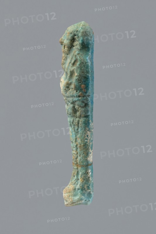 Ushabti, Mummy-shaped figurine funerary offering, wrapped in a shroud with its arms crossed on its chest, holding hoes. Inscription on the front in the form of the letter T. 332–30 BC; Ptolemaic Period, Egypt. Ushabti; height 10 cm, width 2.6 cm., A vertical elongated object against a plain gray background that is predominantly turquoise with areas of darker teal and lighter aqua, showing rough, pitted and irregular texture with patches of white and small rusty brown spots, an indented upper section with subtle raised forms along its length, a slightly wider base that appears rounded and worn, and an overall uneven, weathered surface.