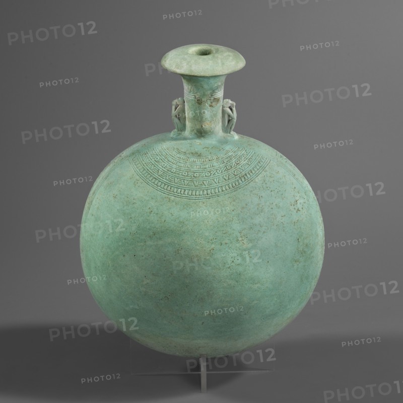New Year's canteen. Flat round faience canteen with a narrow short neck ending in a chipped spout shaped like a papyrus capital. Both sides decorated with modeled figurines of crouching baboons elbows on knees, paws supporting the muzzle. A floral necklace in relief decorates both sides of the neck, and a hieroglyphic text runs along the narrow sides. Materials: faience. Function: vessel. Dimensions: height 17.5 cm +1.5 cm base, width 13.7 cm, weight 0.37 kg. Date: 664–525 BC, Late Period, 26th Dynasty, Egypt. Makers: unknown., A round, flattened vessel with a narrow flared neck and a wide rim sits against a plain gray background, the vessel showing a greenish-blue patina with areas of darker green and subtle brown spots; a decorative band of incised geometric patterns arcs below the neck, and two small sculpted figures flank the neck where it meets the body; the object rests on a clear support with a small vertical element visible at the base.