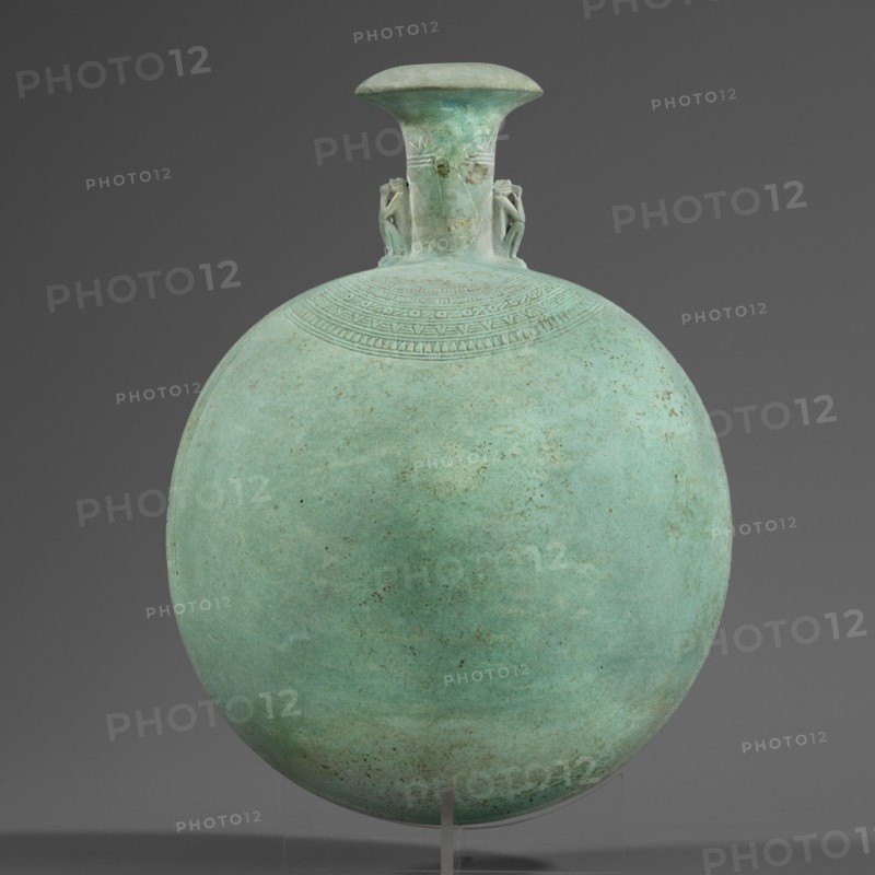 New Year's canteen, Goluchów Greater Poland Voivodeship. Flat round faience canteen with a narrow short neck ending in a chipped spout shaped like a papyrus capital. Decorated on both sides with modeled figurines of crouching baboons, elbows on knees and paws supporting the muzzle. A floral necklace is depicted in relief on both sides of the neck, and a hieroglyphic text runs along the narrow sides. Materials: faience. Function: ritual vessel/bottle. Date: 664–525 BC; Late Period, 26th Dynasty, Egypt. Height 17.5 cm +1.5 cm base; width 13.7 cm; weight 0.37 kg., A round vessel with a flattened spherical body and a short flaring neck set against a plain dark gray background, the vessel displaying a weathered greenish-blue patina with areas of darker green and small brownish spots; a band of incised geometric patterns decorates the upper shoulder just below the neck, and a pair of small sculpted humanlike figures flank the neck where it meets the body; the vessel rests on a clear stand th...