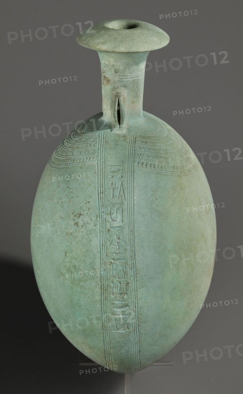 New Year's canteen, Goluchów Greater Poland Voivodeship. Flat round faience canteen with a narrow short neck ending in a chipped spout shaped like a papyrus capital. Decorated on both sides with plastic figurines of crouching baboons, elbows on knees and paws supporting the muzzle. A floral necklace is depicted in relief on both sides of the neck, and a hieroglyphic text runs along the narrow sides. Egyptian, Late Period, 26th Dynasty, 664–525 BC. Vessel; height 17.5 cm plus 1.5 cm base, width 13.7 cm, weight 0.37 kg., A single elongated rounded vessel with a narrow cylindrical neck and flaring rim sits against a neutral gray background; the vessel is predominantly light green with areas of pale turquoise and subtle brown spotting, and it features incised linear decorative bands and a vertical relief strip running down its front, with a small looped handle where the neck meets the shoulder.