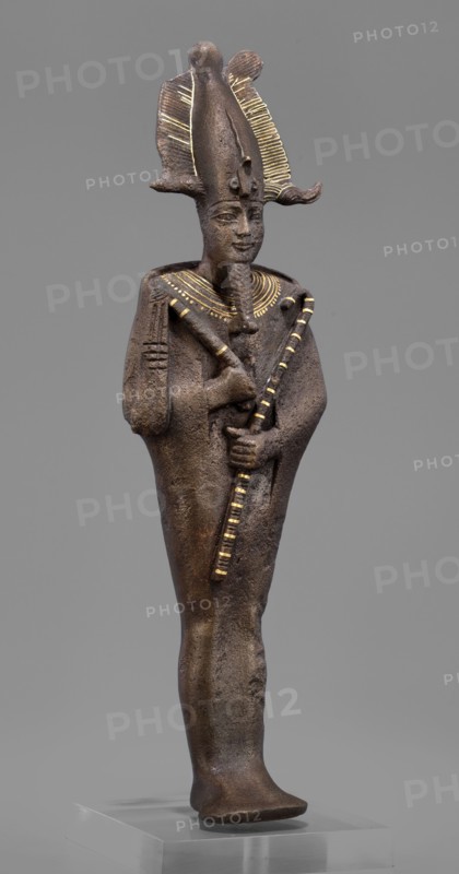 Figurine of the god Osiris; maker, Goluchów Greater Poland Voivodeship; Egyptian deities; crowns; mummy-shaped figurines; male figurines; Egyptian art; Osiris myth.; 664–525 BC, Late Period, 26th Dynasty, Egypt; sculpture; height 29 cm, width 6.8 cm, depth 4 cm., A standing statue of a human figure made of dark brown material with a textured surface, wearing a tall, ornate headdress with decorative ridged elements highlighted in gold, a stylized beard, and a broad collar with gold accents; the figure holds two crossed objects in front of the chest, one of which has alternating gold and dark segments, and the other shows gold detailing; the arms are close to the body and the robe-like garment is smooth and dark brown; the statue stands on a clear rectangular base against a neutral gray background, and visible colors include dark brown, gold, and gray.