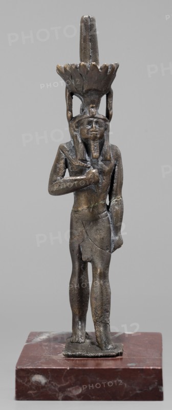 Figurine of the god Nefertum; creator, Egyptian deity Nefertum; Egyptian religion and art; crowns; Late Period, 664–332 BC, Egypt; sculpture; height 12.4 cm, width 3.3 cm, depth 4 cm., A small sculpted figure standing on a rectangular base, rendered in dark brown and bronze tones with areas of lighter brown and greenish patina; the figure wears a headdress with a tall vertical element and a flared, petal-like crown, both in similar dark metallic hues; the figure's facial features, beard, and hair are visible in the same metallic colors, and one arm is bent with the hand placed on the chest while the other arm hangs down; the torso and limbs show textured metal surface and subtle cracks or joins, and the base is a reddish-brown stone with white and lighter brown marbling.