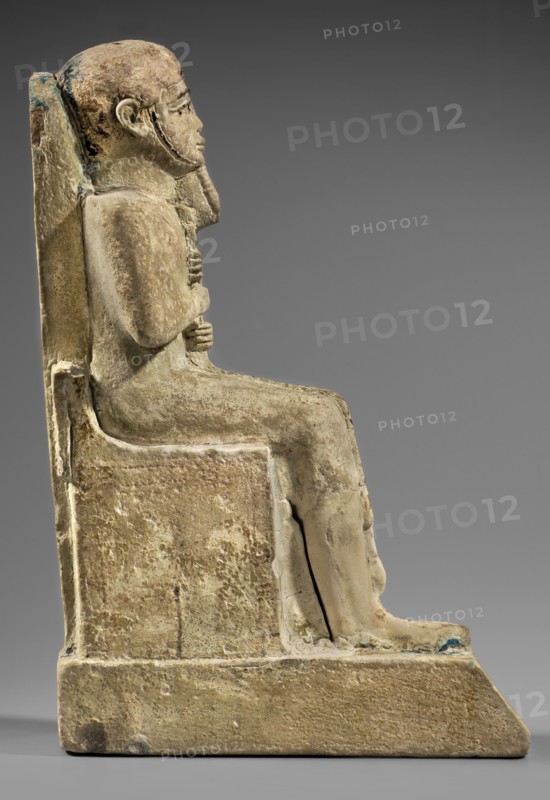 Figurine of the god Ptah sitting on a throne, on a base in the shape of the sign of Maat. He holds a composite scepter in his hands. Materials/attributes: hieroglyphs, ankh, uas, djed pillars, scepters, back pillars., Inscription/Historical details: c. 1550–c. 1295 BC; New Kingdom, 18th Dynasty, Egypt. Object type: figurine. Dimensions: height 29 cm; width 10.5 cm; depth 19 cm; weight 2.736 kg., A stone sculpture of a seated human figure viewed in profile, positioned on a rectangular block-like throne with a high straight back; the figure has a rounded head, a short stylized beard, and hands crossed and resting on the chest, with legs bent at the knees and feet extending forward to a sloped platform. The surface shows rough weathered texture and areas of wear, with overall beige and tan tones, along with patches of darker brown and faint greenish-blue discoloration in places. The background is a smooth neutral gray gradient.