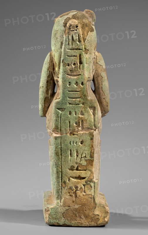 Figurine of the goddess Uadjit with the head of a lioness; Uadjit mythology; Egyptian deities; made of material not specified; hieroglyphs; back pillars; throne; lotuses; inscription Scen.Gal.Eg., 664; date: 30 BC – Late Period to Ptolemaic Period; origin: Egypt; figurine; height 22 cm, width 11.5 cm, depth 7 cm, weight 1.024 kg; maker, attributed period ca. 30 BC., A small carved figurine photographed from the back, showing a standing anthropoid form on a rectangular base; the surface is worn and pitted with a mottled green and brown coloration, and areas of lighter beige where material has flaked away; the figure has two rounded shoulder/arm forms projecting at its sides and a central vertical panel of shallow incised shapes; the base is squared with irregular edges and the overall object shows signs of age with rough texture and patches of discoloration against a neutral gray background.