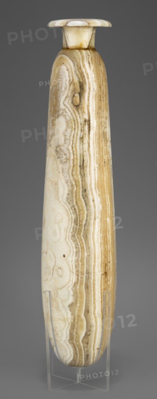 Alabastron, Slender alabastron with a distinct short cylindrical neck and a wide flat rim, provided with two tiny handles in the form of suppositories; stone vessel for oils and ointments, Egyptian art, Late Period, 664–332 BC; vessel alabastron, height 32 cm +1.5 cm base, width 5 cm, weight 1.11 kg, A tall, narrow, cylindrical vessel with a flared rim displayed upright on a clear stand, showing layered banding and swirling patterns in shades of beige, cream, tan, and light brown, with subtle darker brown streaks and small spots; the surface appears smooth and polished with natural banded striations running vertically along the body and concentric swirls visible in places, set against a plain gray background.