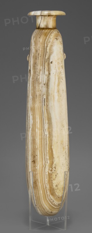Alabastron, Slender alabastron with a distinct short cylindrical neck and a wide flat rim, provided with two tiny handles in the form of suppositories. Stone vessel for oils and ointments alabastron; Egyptian art; Late Period, 664–332 BC. Height 32 cm +1.5 cm base, width 5 cm, weight 1.11 kg., A tall, elongated cylindrical object with a rounded top and a small flared lip, displayed upright on a clear stand; its surface shows layered, banded patterns in shades of beige, cream, tan, and light brown with areas of mottled discoloration and darker speckles, and the background is a solid medium gray.